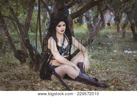 Beautiful Brunette Girl In A Suit With Horns Sitting Under A Tree.