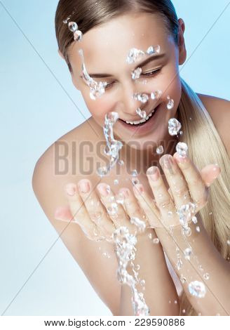 Portrait of a young beautiful woman cleaning her face with fresh cold water, isolated on blue background, morning freshness, removing makeup, health and beauty care concept