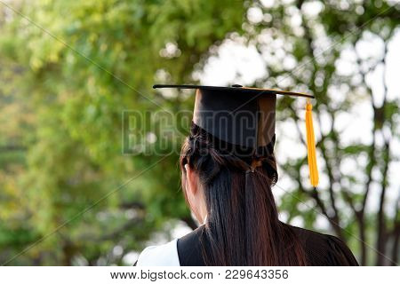 Behind Photo Of University Graduate Wears Gown And Black Cap, Yellow Ribbon And Blur Bokhe Backgroun