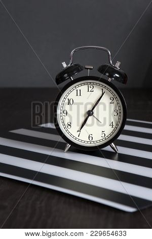 Black Alarm Clock On A Black And White Striped Napkin Showing 7 O'clock On A Bedside Table