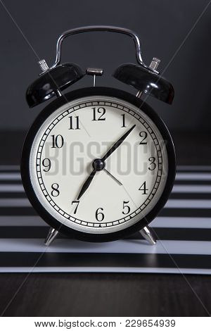 Black Alarm Clock On A Black And White Striped Napkin Showing 7 O'clock On A Bedside Table