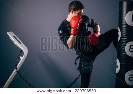 Fit Man In Boxing Gloves Training Knee Kick. Kickboxer Wearing Ems Suit Pounching Bag