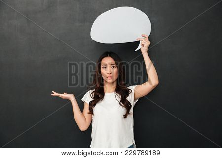Confused asian woman in t-shirt holding copyspace on the palm and looking at the camera while holdind blank speech bubble over black background