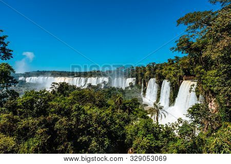 A Green And Lush Rainforest Is Overlooking The Huge Iguazu Falls On A Clear And Sunny Day.