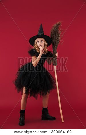 Image of serious little girl witch in carnival halloween costume isolated over red wall background with broom showing silence gesure.