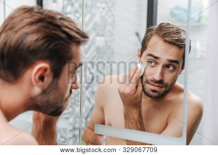 Handsome young shirtless man applying aftershave lotion with a cotton pad while standing at the bathroom mirror