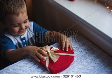 Happy Child Boy Holding Red Heart Shaped Gift Box With Bow Near Window In Daylight. Valentine Day Pr
