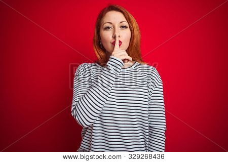 Young redhead woman wearing strapes navy shirt standing over red isolated background asking to be quiet with finger on lips. Silence and secret concept.