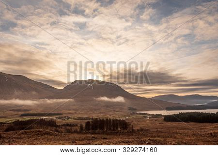 A View Of The Wintery Grass Fields In The Scottish Highlands As The Sun Sets Over A Mountain On A Cl