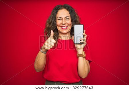Middle age senior woman showing screen of smartphone over red isolated background happy with big smile doing ok sign, thumb up with fingers, excellent sign