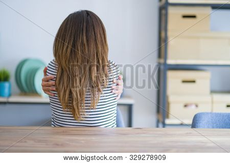 Middle age senior woman sitting at the table at home Hugging oneself happy and positive from backwards. Self love and self care