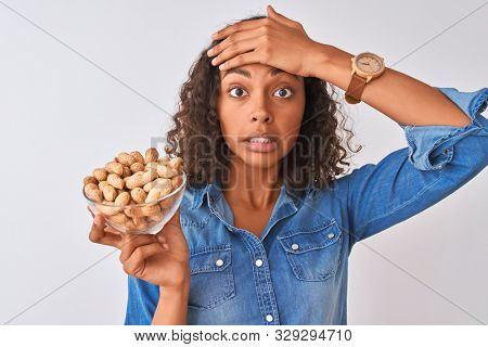Young brazilian woman holding bowl with peanuts standing over isolated white background stressed with hand on head, shocked with shame and surprise face, angry and frustrated. Fear and upset