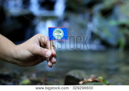 Flag Of Belize On Wooden Stick. Afro American Holds National Athem Near Waterfall On Stream In Autum