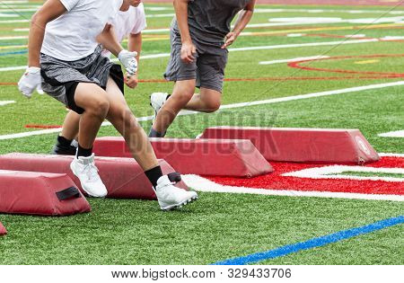 Three Football Players Are Running Between Red Barriers On A Turf Field During Summer Camp Training.