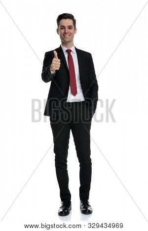 Cheerful businessman and giving a thumbs up and holding hsi hand in his pocket while wearing a black suit and red tie, standing on white studio background