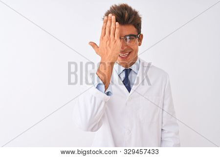 Young handsome sciencist man wearing glasses and coat over isolated white background covering one eye with hand, confident smile on face and surprise emotion.