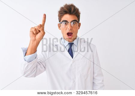 Young handsome sciencist man wearing glasses and coat over isolated white background pointing finger up with successful idea. Exited and happy. Number one.
