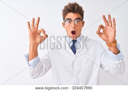 Young handsome sciencist man wearing glasses and coat over isolated white background looking surprised and shocked doing ok approval symbol with fingers. Crazy expression