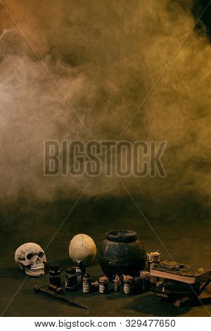 Steaming Pot Against A Black Studio Background. There Are Jars With Ingredients For A Potion, Skulls