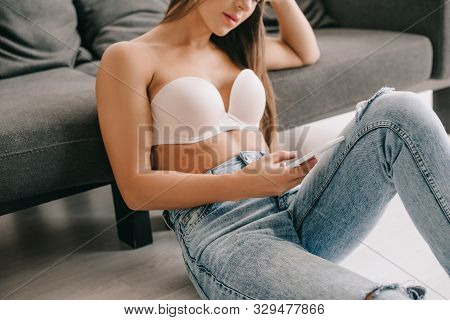 Cropped View Of Woman In White Bra Using Smartphone While Sitting On Floor Near Sofa