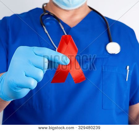 Doctor In Blue Uniform And Sterile Latex Gloves Holds A Red Ribbon-symbol Of The Fight Against Disea