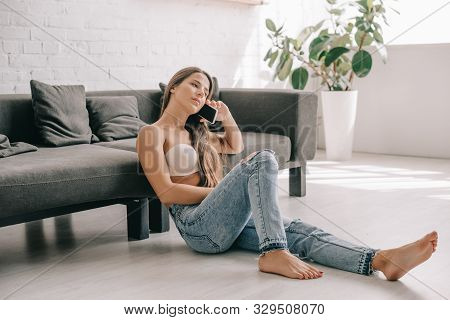 Beautiful Girl In White Bra And Jeans Talking On Smartphone While Sitting On Floor Near Sofa
