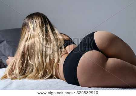 A Blonde With Long Hair In Black Underwear Is Lying On The Bed. Rear View Of A Girl In Thong Sleepin