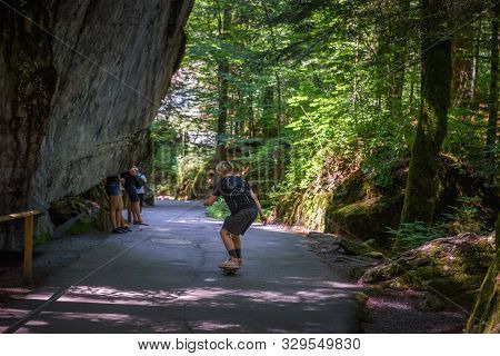 Young Skater Riding A Skateboard On A Road Towards The Blausee Lake In Switzerland, Surrounded By De