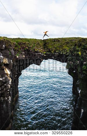 Tourist Jumps Over A Natural Rock Bridge Connecting Basalt Cliffs In Arnarstapi, Iceland, With Atlan