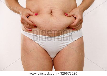 An Elderly Woman In White Panties Shows The Folds On Her Abdomen On A White Isolated Background.