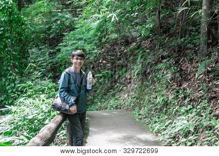 Asian Male Tourist With Camera Sling Bag Holding A Bottle Of Water On Foot Path Traveling Into The W
