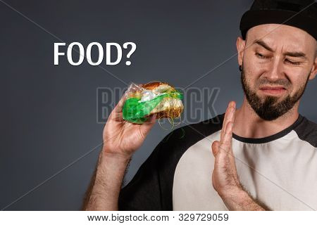 The Concept Of Fast Food And Ecology. A Man In A Cap And A Beard, Holding A Hamburger With A Plastic