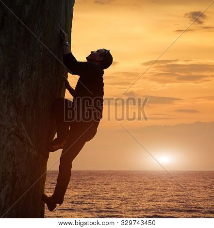 Side View Of Man In Black Climbing On Rock Without Safety Harness Over Sea In Twilight. Recreational