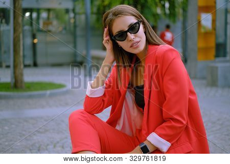 Blonde Girl In A Red Lady-type Pantsuit And Black Top, Watch, Ring, Pendant And Sunglasses Is Posing