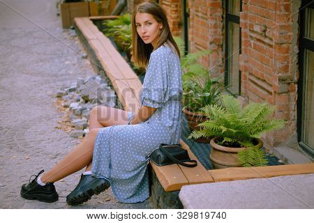 Blonde Girl In Long Blue Dress And Small Black Handbag On Her Shoulder Is Posing Sitting Sideways On