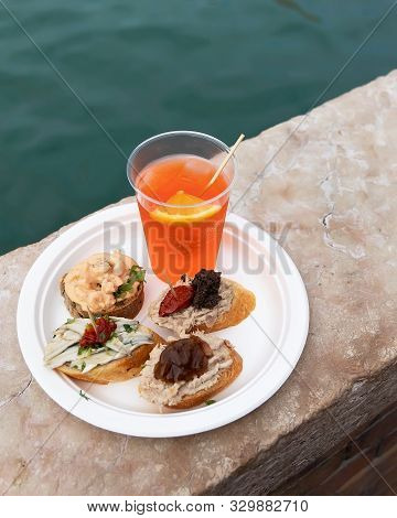 Traditional Venetian Food And Drink Cicchetti And Aperol Spritz With Turquoise Green Water Of Canal.