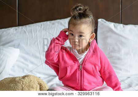 Cute Asian Baby Girl Wearing A Pink Jacket Sitting On The Bed.