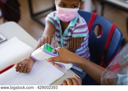 Mid section of female health worker measuring temperature of a girl at elementary school. education back to school health safety during covid19 coronavirus pandemic.