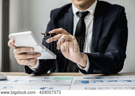A Man With A Pen Pointing To A White Calculator And Documents Are Placed On The Table, A Businessman