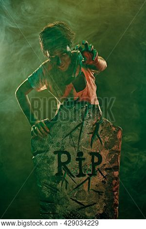 Horrible Halloween. Scary zombie boy teenager stands next to the headstone and gloats in the cemetery. Copy space.