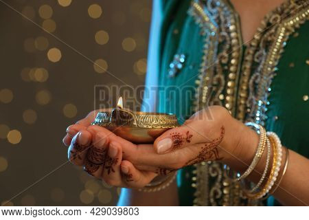 Woman Holding Lit Diya Lamp On Brown Background With Blurred Lights Closeup View. Diwali Holiday