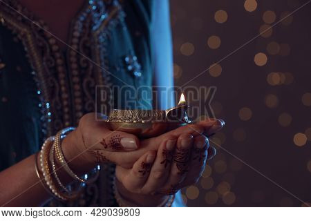 Closeup View Of Woman Holding Lit Diya Lamp On Brown Background With Blurred Lights, Space For Text.