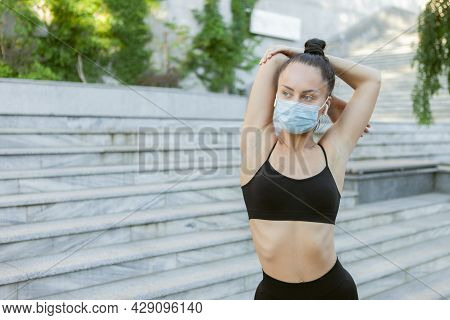Young Slim Woman In Medical Protective Mask Doing Misk Stretching Before Training Outdoors. Sports D