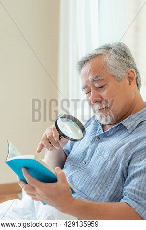 Asian Senior Old Man Longsighted , Farsighted , Reading A Book With A Magnifying Glass On White Bedr