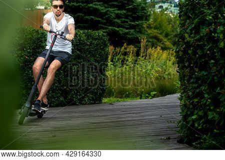 Man Riding Electric Scooter At Beautiful Park Landscape. Man Is On Foreground, Modern Park Is On Bac