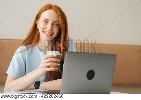 Close-up Face Of Happy Young Beautiful Woman Holding Cup With Hot Coffee Sitting At Desk With Laptop