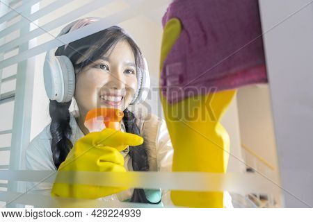 An Asian Female Maid Is Cleaning The House By Wiping The Glass, Happily Wearing Music Headphones. Us