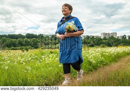 Grandmother Is Walking Along A Path In A Field Of Flowers. An Elderly Woman Walks In Nature.