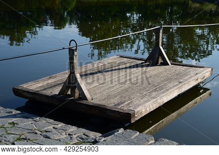 A Wooden Raft Is Tied To A Taut Rope. Self-service Ferry Across The River, Pond. People Get On And P