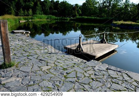 A Wooden Raft Is Tied To A Taut Rope. Self-service Ferry Across The River, Pond. People Get On And P